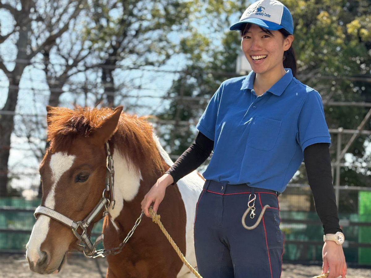 ポニーのリードを持ち、笑顔で立つスタッフの女性。「Harmony Center」のキャップを着用。