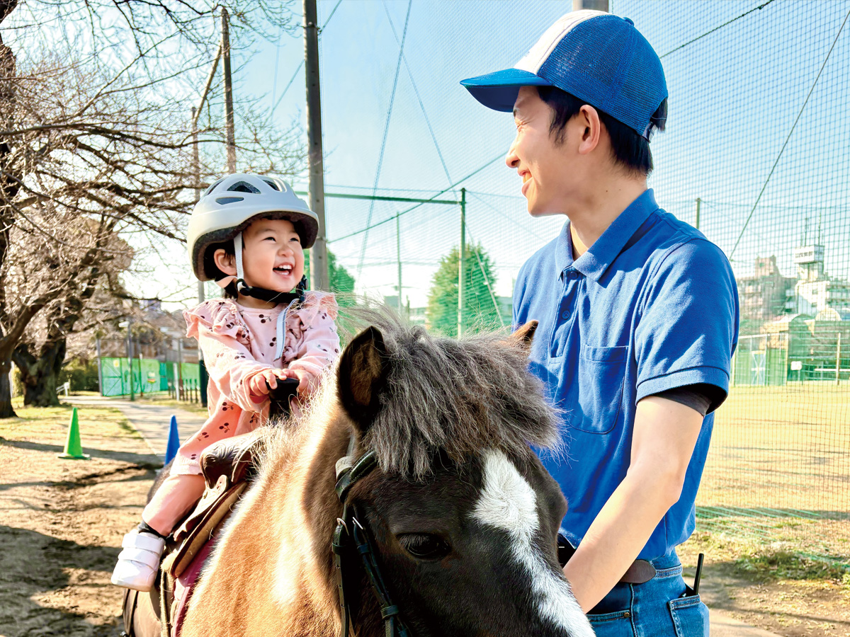 ポニーに乗って笑顔の幼い子どもと、青いポロシャツ姿で寄り添うスタッフの男性。