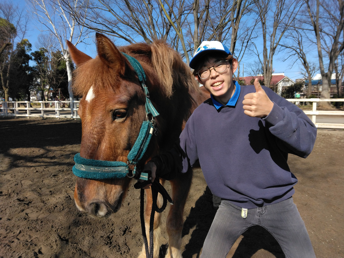 馬のそばに立ち、サムズアップポーズをするスタッフの男性。青空と木々が広がる馬場にて。